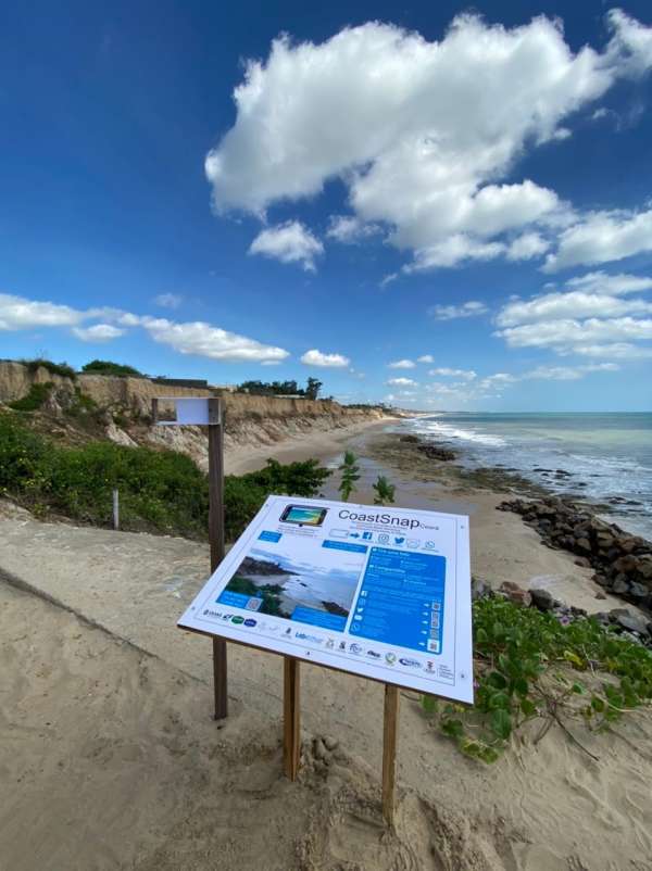 A imagem mostra a estação piloto, na praia do Pacheco, no Ceará. A imagem mostra o céu azul com algumas nuvens e o mar. Em primeiro plano está a estação, semelhante a uma placa na horizontal, com o nome do Projeto CoastSnap Ceará, sobre estrutura simples de madeira.