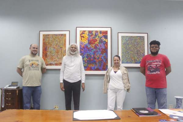 A imagem mostra quatro pessoas em pé, lado a lado, posando em frente a três quadros coloridos pendurados na parede. As pinturas, que são abstratas e ricas em cores, parecem ser compostas de formas pontilhadas e padrões complexos. Cada obra tem moldura branca.
