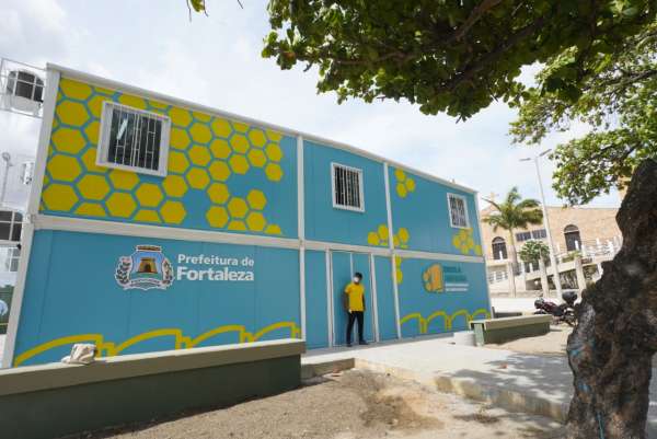 Foto da fachada de um prédio azul claro, de um andar, com alguns losangos amarelos. Na parede consta a logomarca do projeto Escola Areninha e, do outro lado, a logomarca da Prefeitura de Fortaleza. A imagem foi feita durante o dia, com céu claro. Há, ainda, um jovem porteiro na entrada do prédio.