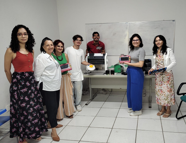 #PraTodoMundoVer: Na foto, em semicírculo, de pé, membros do NAAI, Agin e Incubadora. Professora Renata (NAAI) e Beatriz (Agin) seguram a lupa, Gustavo o mouse acessível e Juliana (Incubadora) a linha braile. Ao centro, mesa com ferramentas assistivas. 